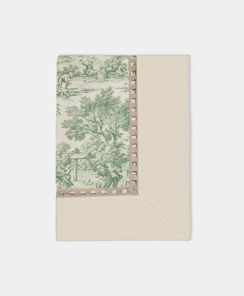 A folded cream-colored napkin with a detailed green pastoral toile print on one section, bordered by a decorative beige ribbon trim.