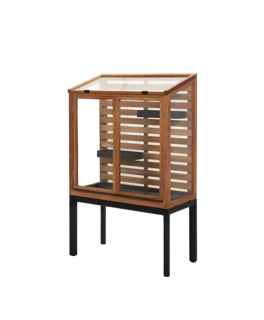 A small wooden greenhouse with a slanted glass roof, glass door, and horizontal wooden slats, standing on tall black legs.