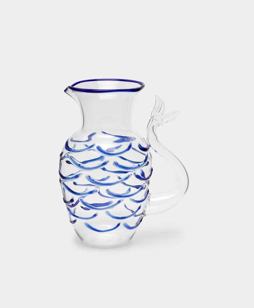 Clear glass pitcher with a handle shaped like a fish tail, decorated with blue wave patterns and a blue rim around the spout. The background is plain light gray.
