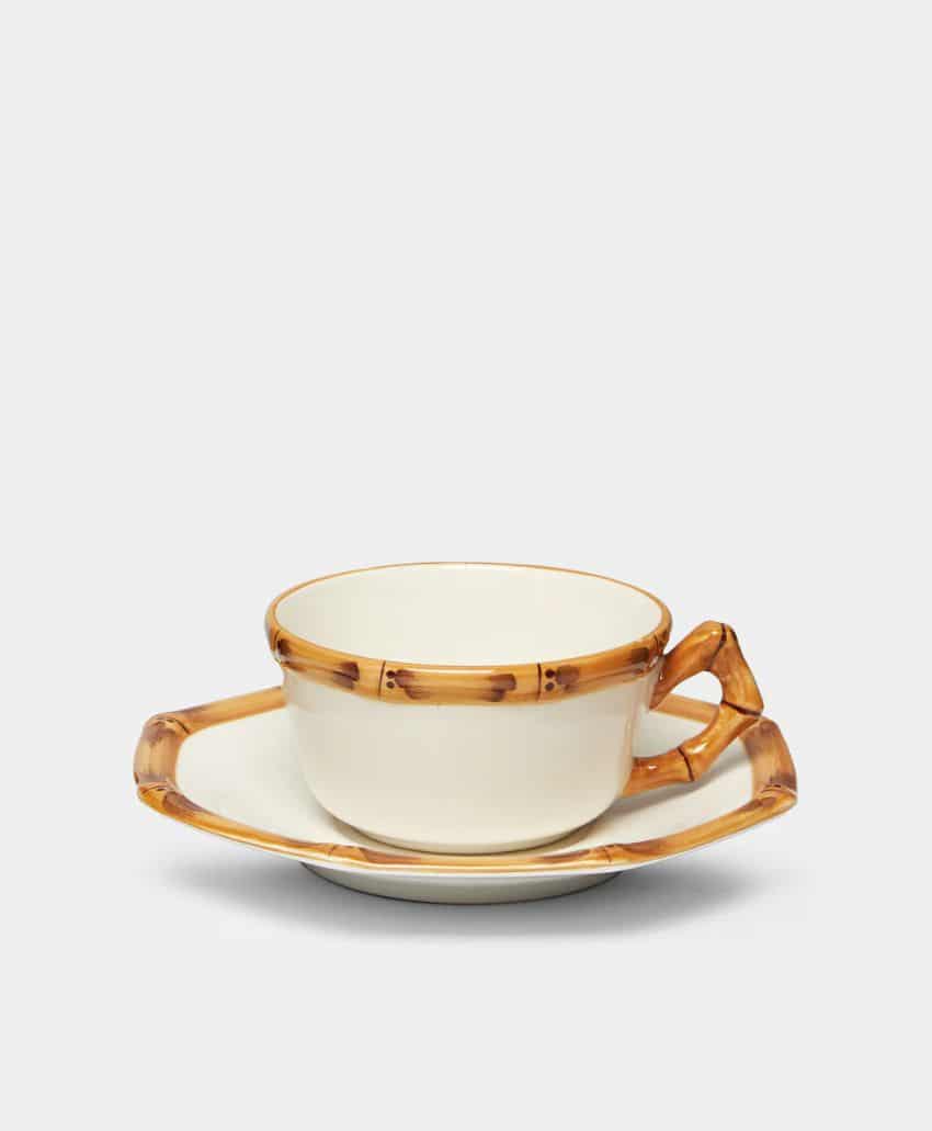 Cream-colored cup and saucer set with a brown, bamboo-style rim and handle, placed against a plain light gray background.
