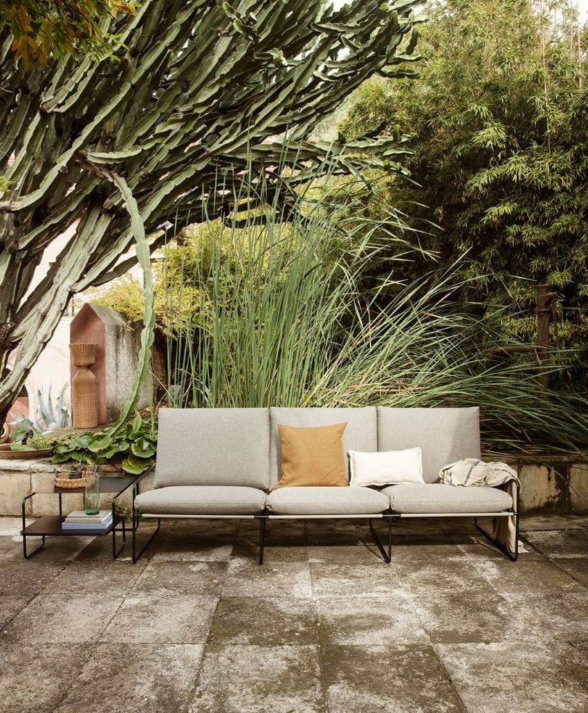 A modern outdoor sofa with gray cushions and a tan accent pillow sits on a stone patio surrounded by lush green plants, including tall grasses, bamboo, and a large cactus. A small side table holds books and a drink.