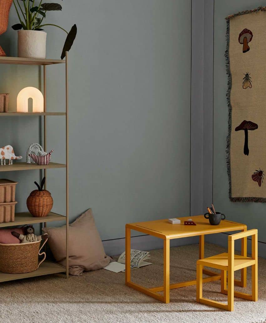 A cozy room with a yellow childrens table and chair, a beige shelf with toys, books, and plants, a cushion on the floor, and a wall hanging featuring mushrooms. The decor is minimalist and warm.