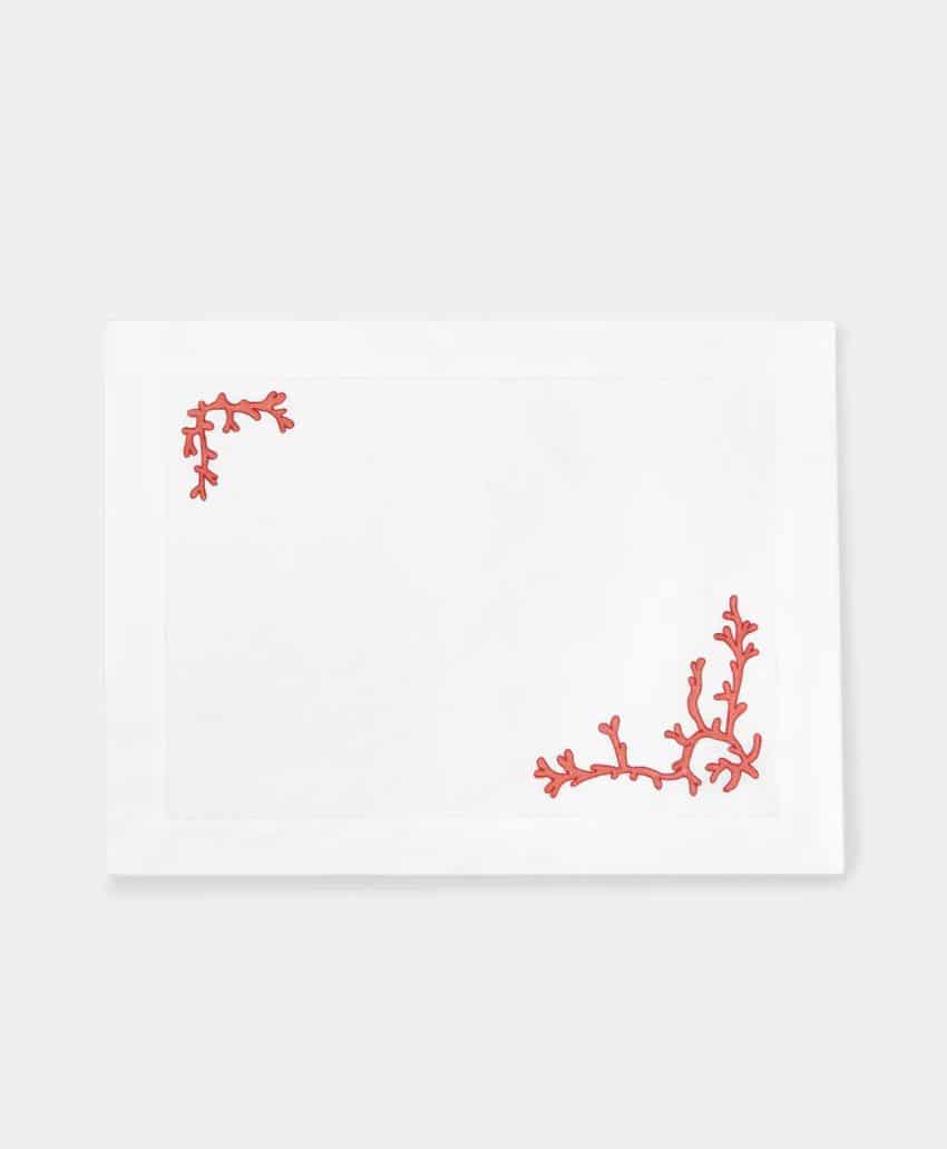 White rectangular placemat with two red coral-shaped embroidered designs in the upper left and lower right corners, set on a plain light gray background.