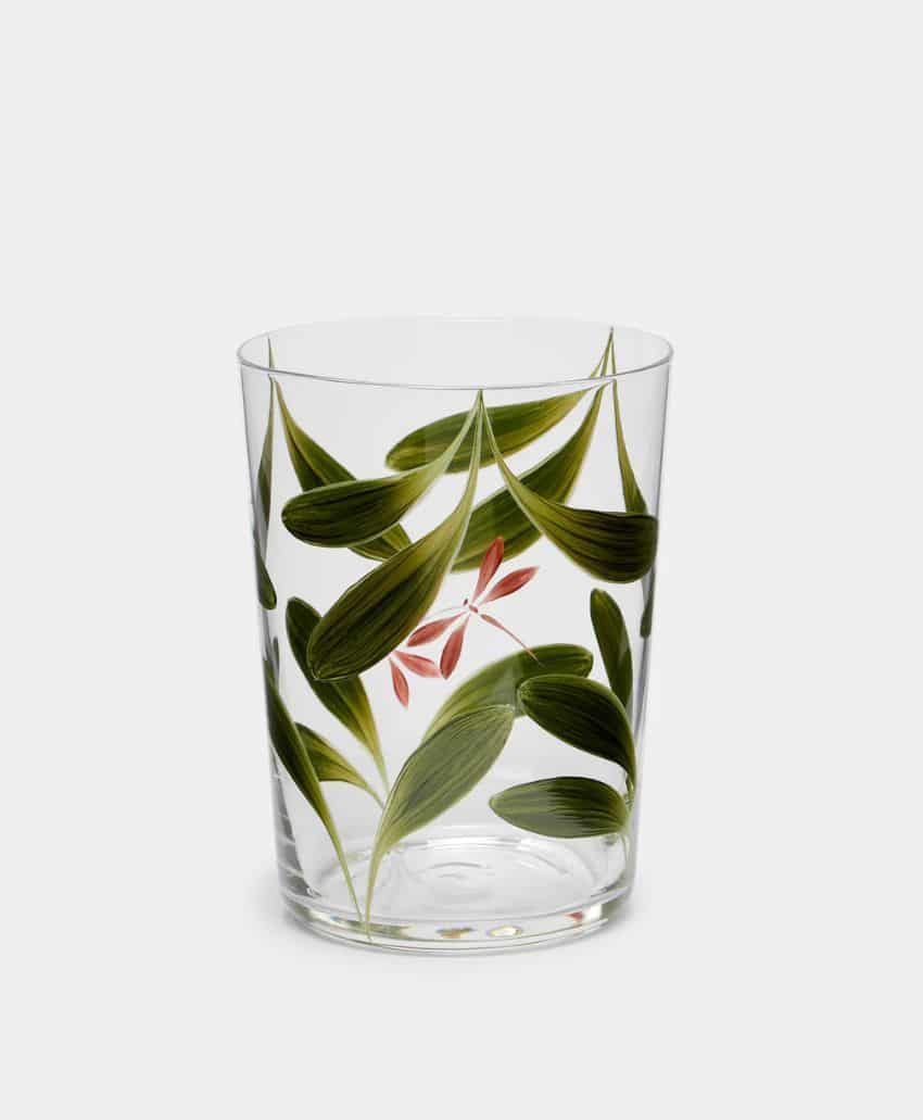 A clear drinking glass decorated with green leaves and a small pink flower, set against a plain light gray background.