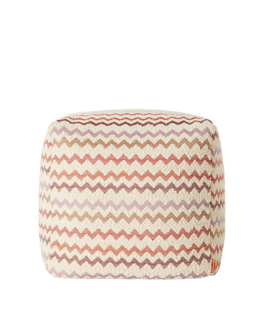 A fabric pouf with a square shape, featuring a zigzag pattern in muted shades of pink, orange, beige, and purple. The background is plain white.