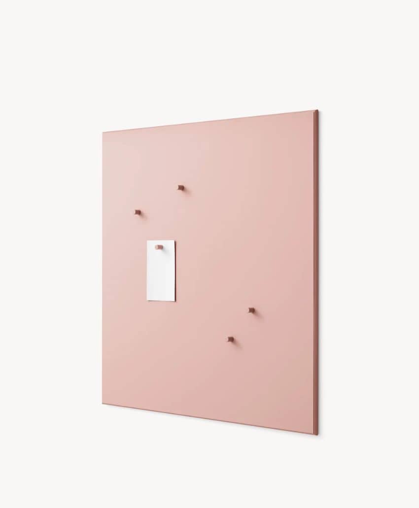 A large, light pink bulletin board hangs on a white wall, with five small pins and a single white note attached near the center. The board has a minimalist, modern design.
