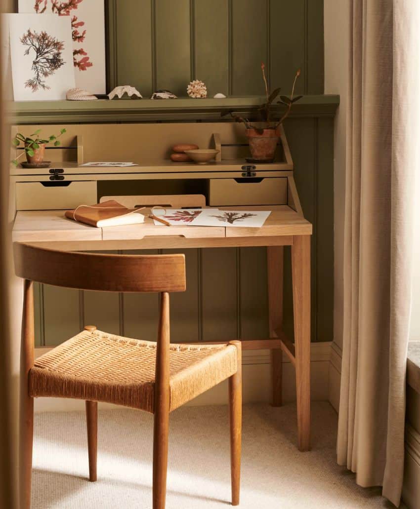 A small wooden desk with papers, books, and potted plants sits in front of a green paneled wall. Above, there are seashells and botanical prints. A wooden chair with a woven seat is in front of the desk by a window with beige curtains.