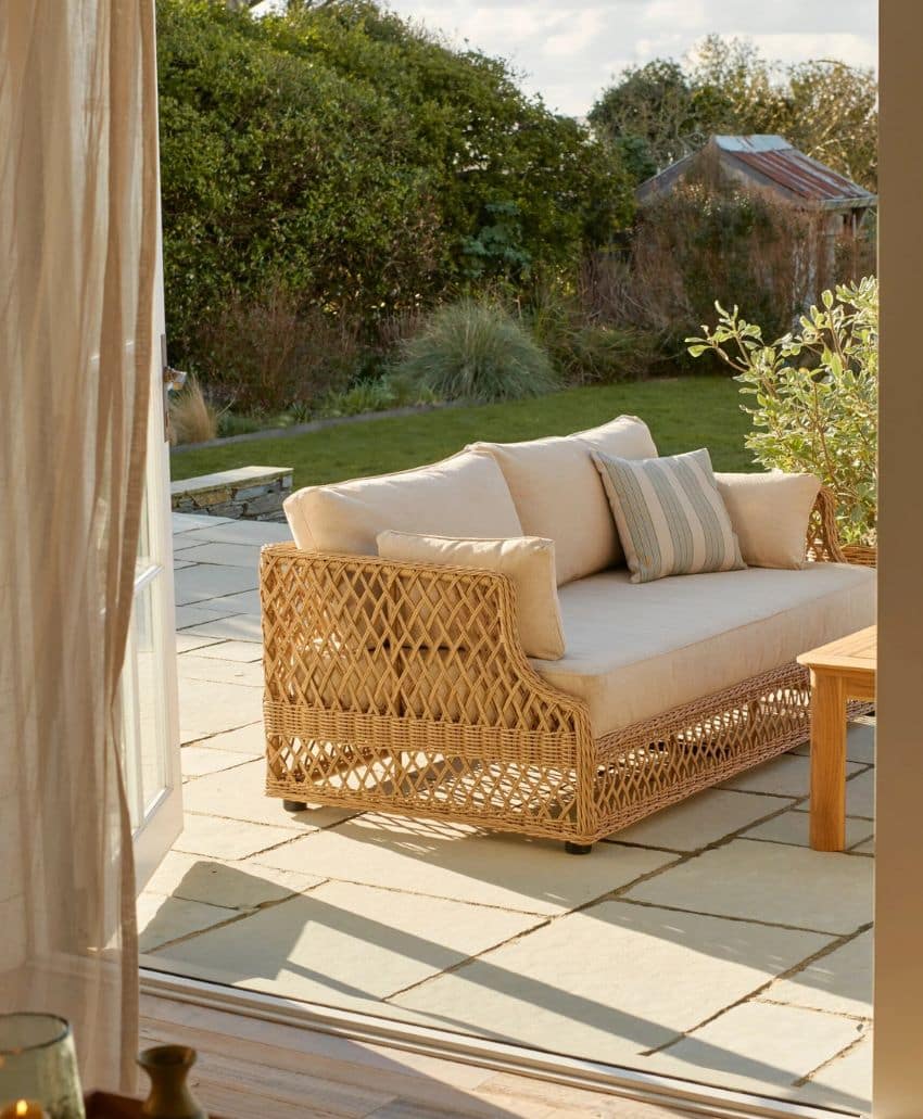 A wicker outdoor sofa with beige cushions and a striped pillow sits on a stone patio, seen through an open glass door, with a garden and trees in the background.