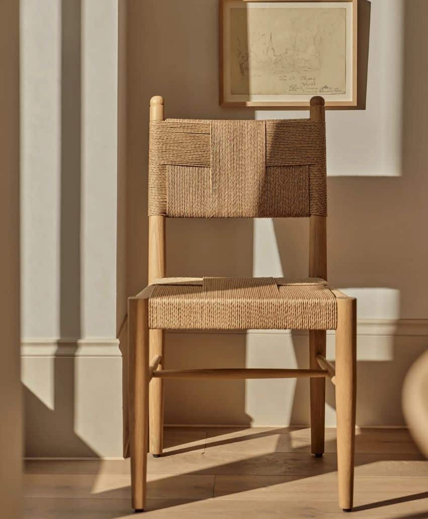 A wooden chair with a woven seat and backrest sits in sunlight against a beige wall, with framed artwork hanging above it. Shadows and warm light create a calm, minimalist atmosphere.