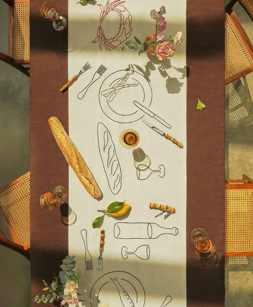 A table set for a meal with a brown runner, baguettes, wine glasses, corkscrews, plates, and floral centerpieces. The runner has black line drawings of tableware and food. Four wooden chairs are visible.