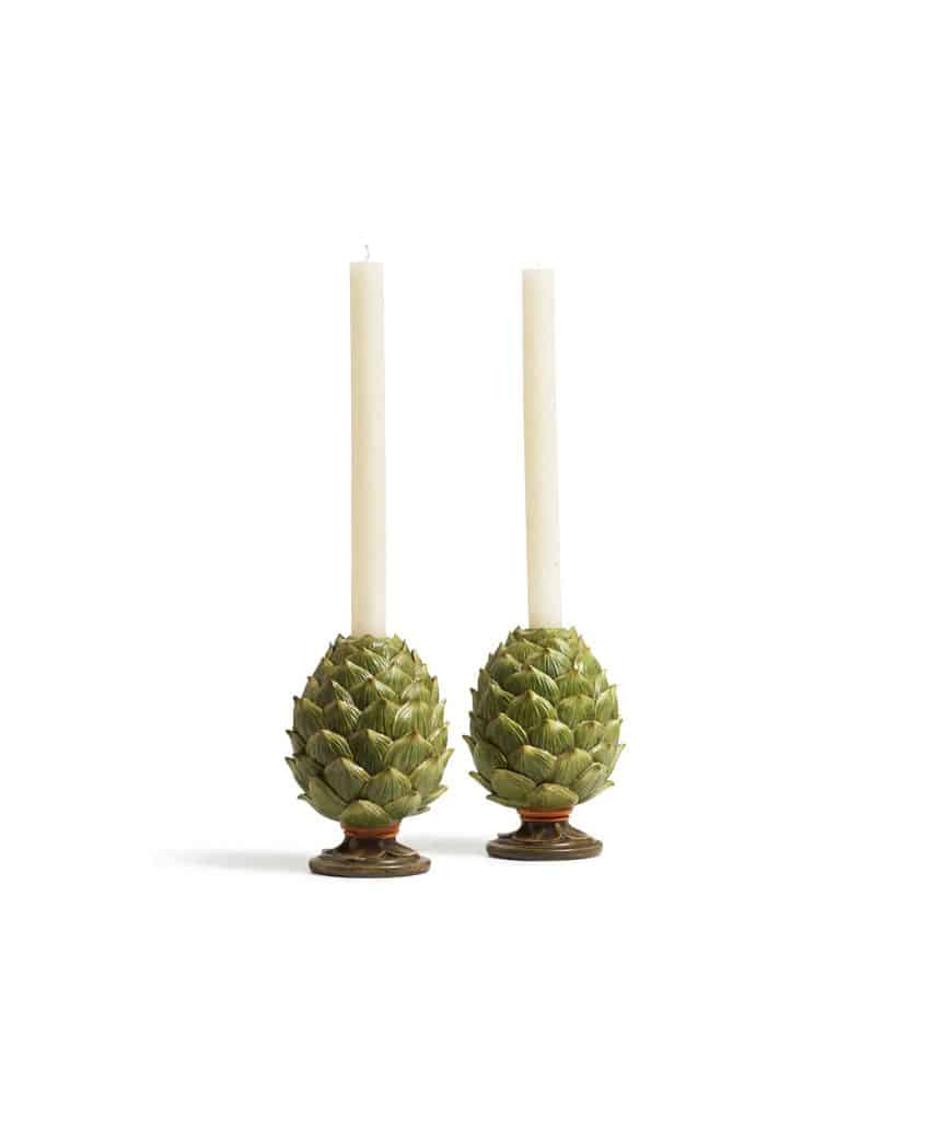 Two tall, white taper candles are placed in holders shaped like green artichokes with round bases, displayed against a plain white background.