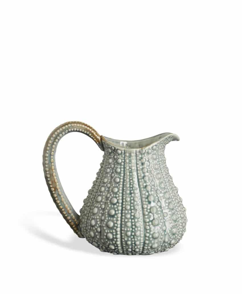 A ceramic pitcher with a textured, sea urchin-inspired design, featuring raised bumps and a curved handle, in soft grey and beige tones, set against a plain white background.