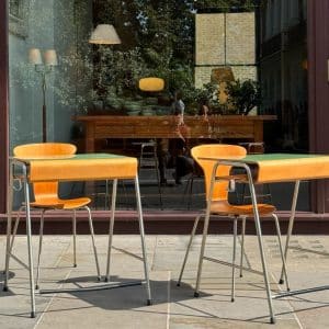 Two wooden chairs with metal legs and tables sit on a stone patio outside a cafe. The glass window behind them reflects trees, a streetlamp, and people, showing a cozy interior with more furniture and lamps.