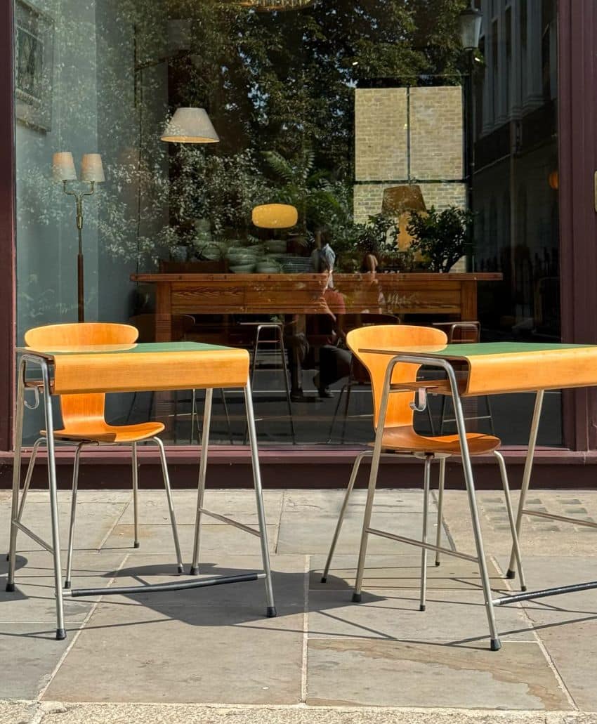 Two wooden chairs with metal legs and tables sit on a stone patio outside a cafe. The glass window behind them reflects trees, a streetlamp, and people, showing a cozy interior with more furniture and lamps.