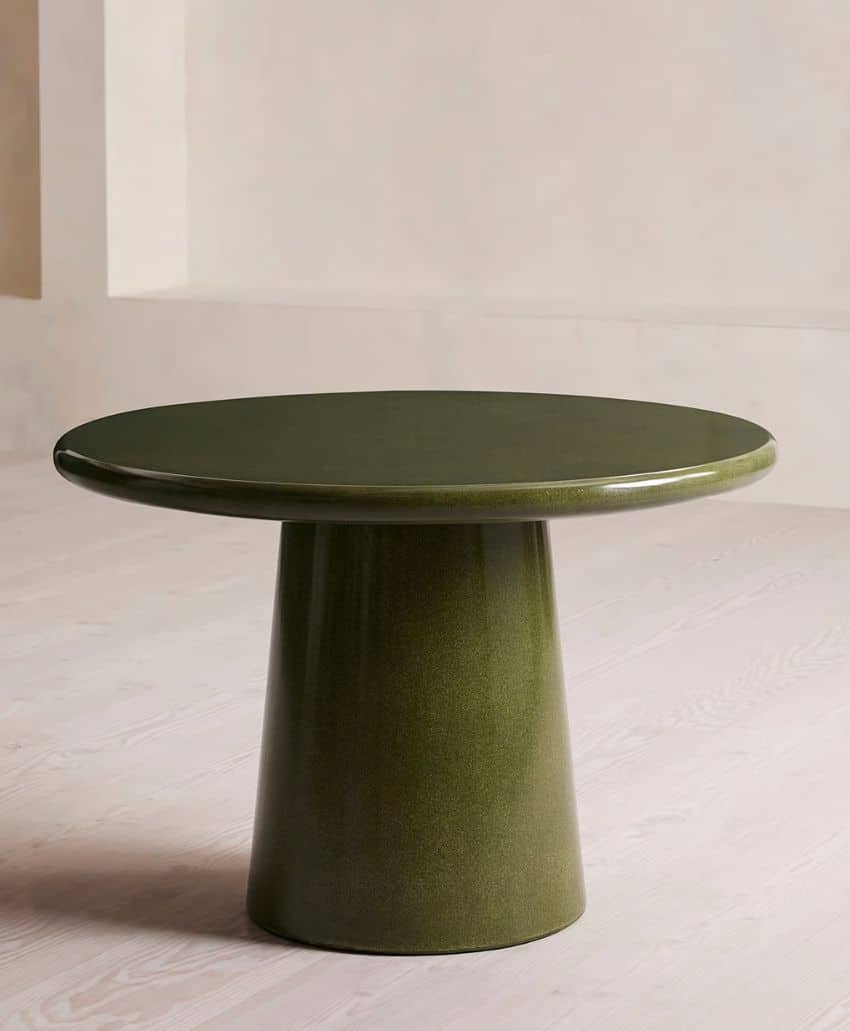 A round, olive green table with a smooth, glossy surface and a wide, tapered pedestal base, placed on a light-colored wooden floor against a neutral wall.