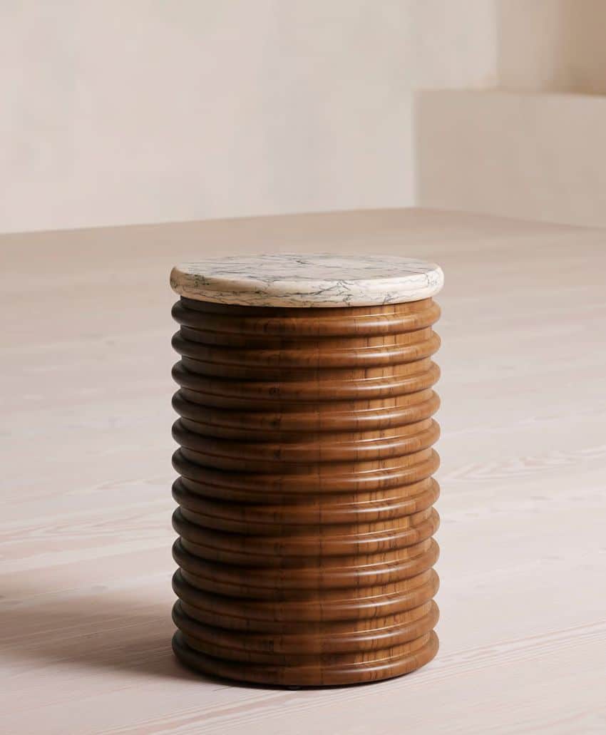 A small, round side table with a ribbed wooden cylindrical base and a smooth, white marble top, placed on a light-colored wooden floor in a minimalistic room.