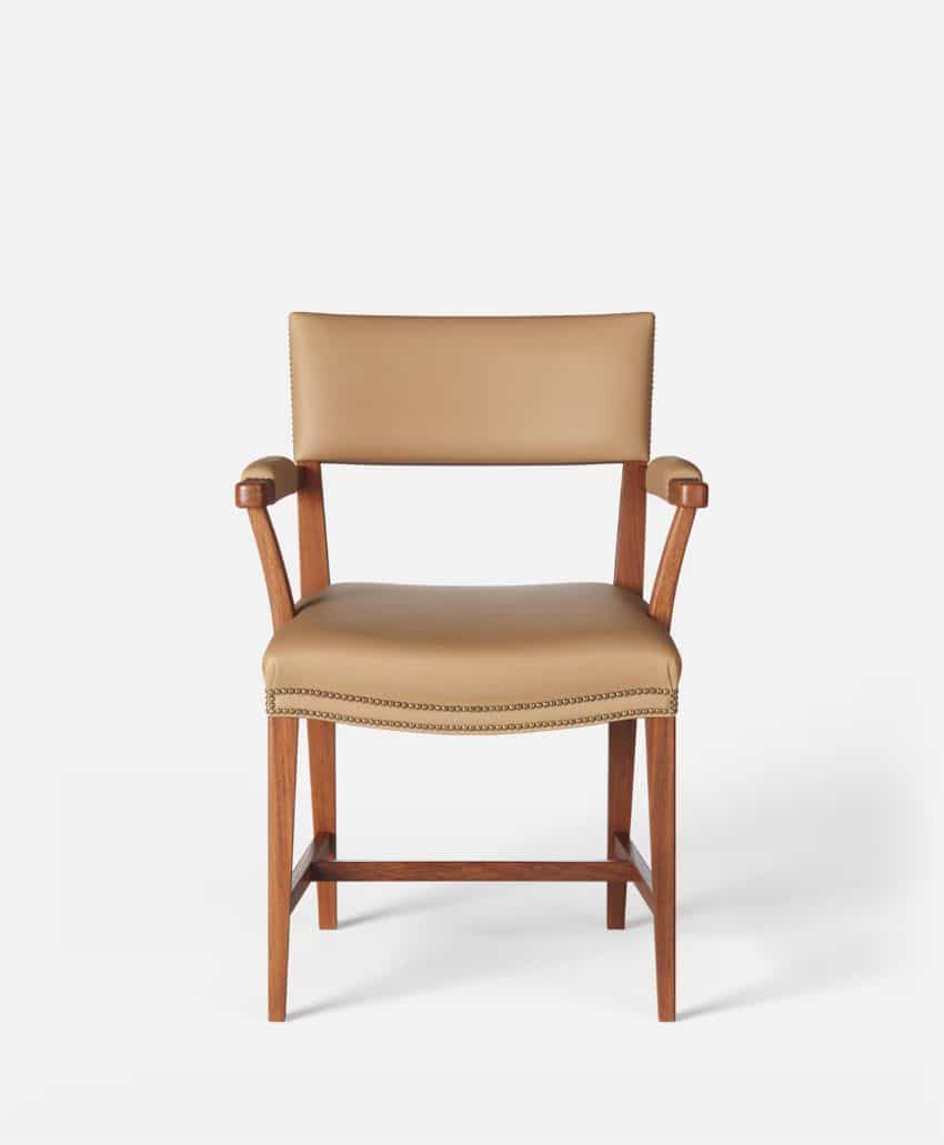 A wooden chair with tan upholstered seat, backrest, and padded armrests, featuring nailhead trim along the edges, set against a plain white background.