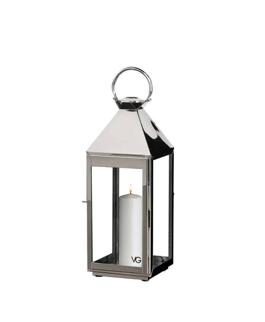 A polished silver metal lantern with glass panels, a round handle on top, and a lit white pillar candle inside. The letters VG are visible on the bottom corner of the lantern.