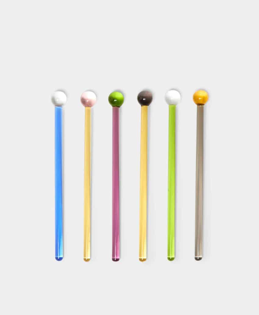 Six glass stirring sticks with round tops, each in different colors: blue, yellow, pink, green, clear, and amber, arranged vertically in a row on a light background.