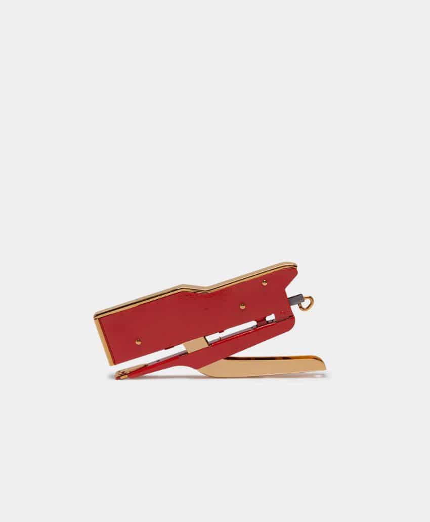 A red and gold handheld stapler is shown in a closed position against a plain light gray background.