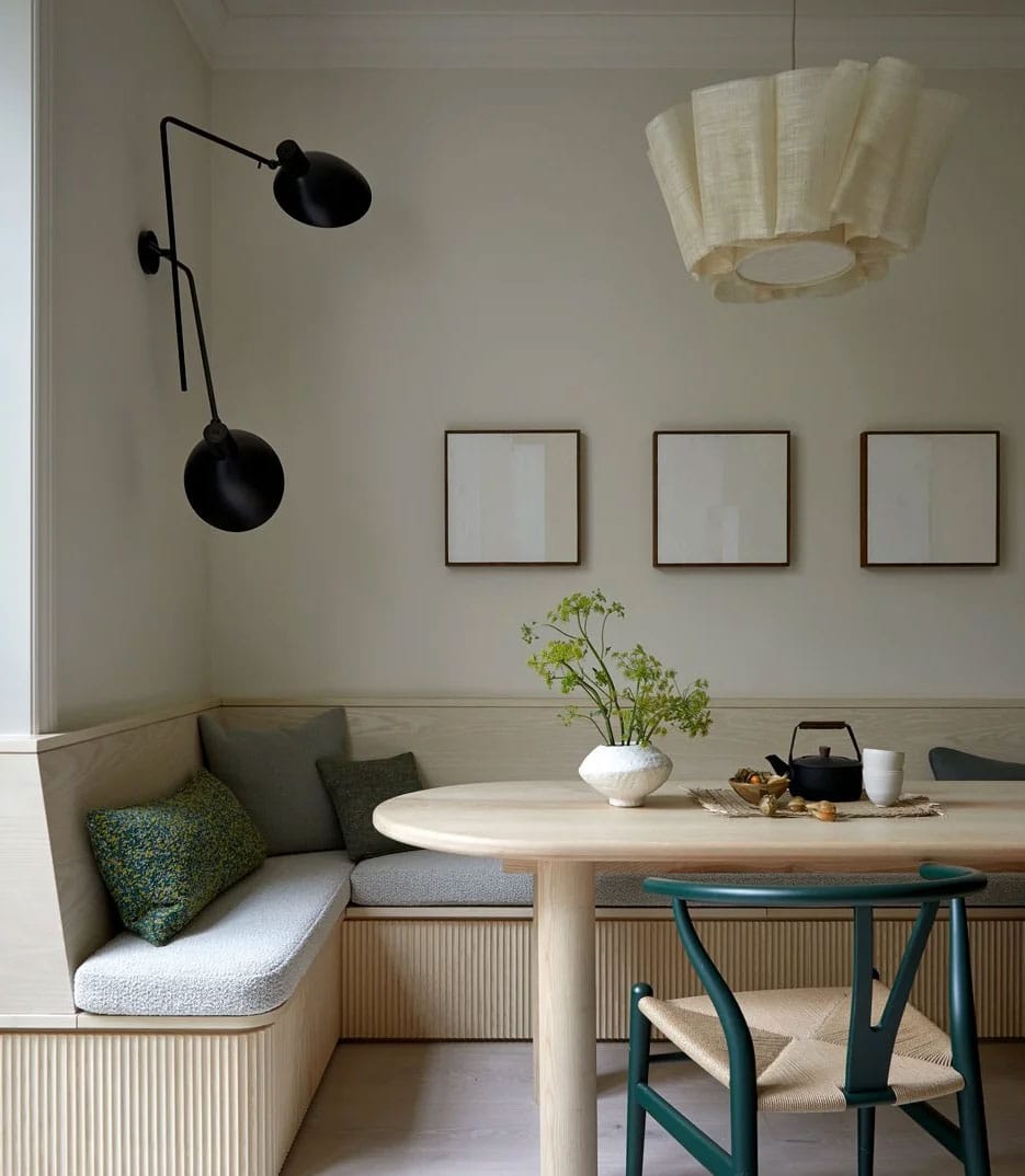 A cozy, modern dining nook with a round wooden table, a built-in bench with green cushions, a green chair, a white vase with greenery, minimalist wall art, a sculptural pendant lamp, and a black wall sconce.
