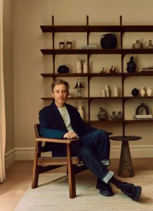 A man in a dark suit sits in a wooden chair beside a small round table. Behind him, a wall-mounted shelf displays various vases, pottery, and decor items. The setting is modern and warmly lit.