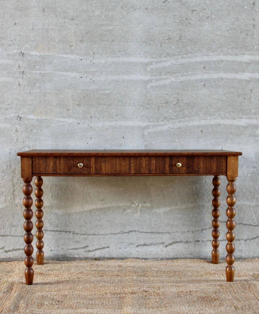 A wooden table with a rectangular top, single central drawer with two round knobs, and four legs featuring a series of rounded, spindle-like segments, placed on a textured rug against a gray concrete wall.