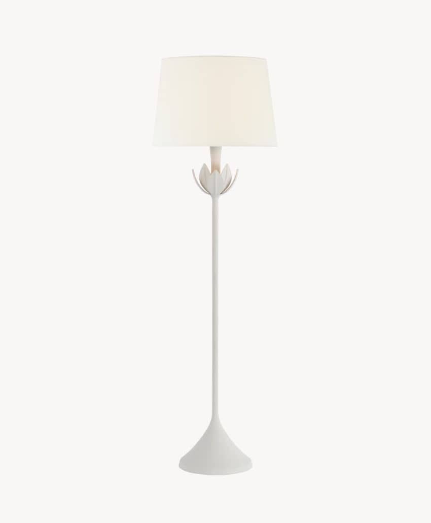 A modern white floor lamp with a wide, round base, a slender stem, and a white lampshade. The lamp features a decorative flower-like detail just below the lampshade.