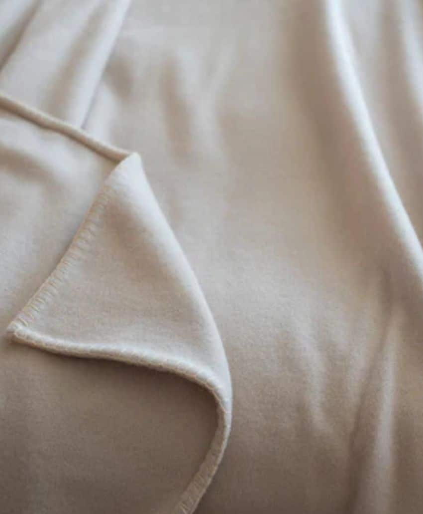 A close-up view of a soft, light beige fabric blanket with smooth texture, slightly folded at one corner and gently draped, creating soft, flowing lines and shadows.