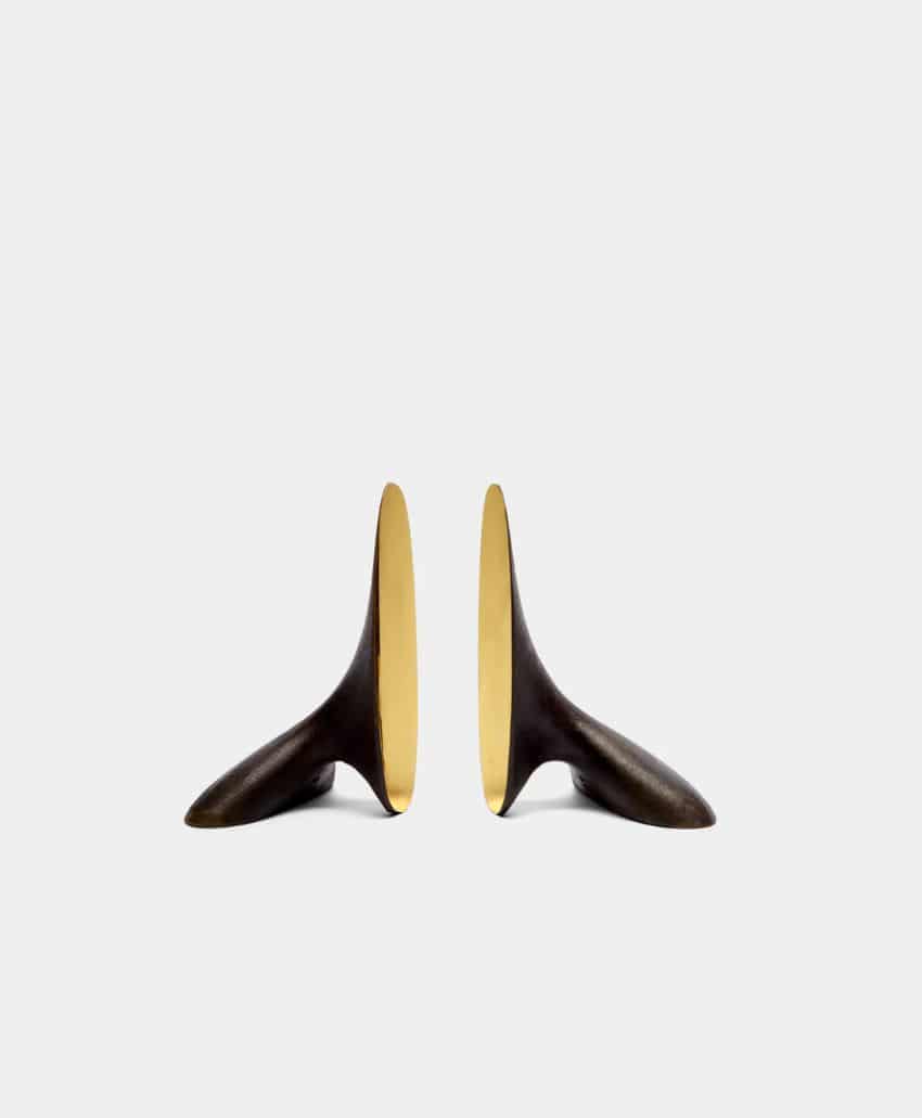 Two sculptural, abstract bookends with curved, smooth shapes. The exterior is dark bronze, while the inner surfaces are gold, creating a striking contrast against a plain light gray background.