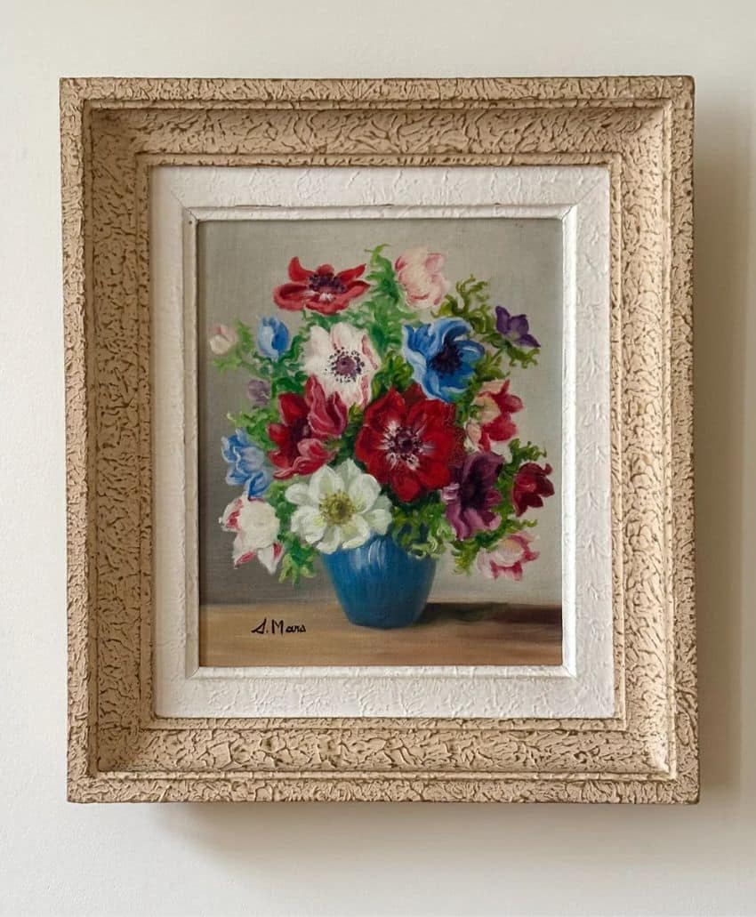 A framed painting of a colorful bouquet of flowers in a blue vase, displayed against a light background. The ornate, textured frame is cream-colored with a white inner border.