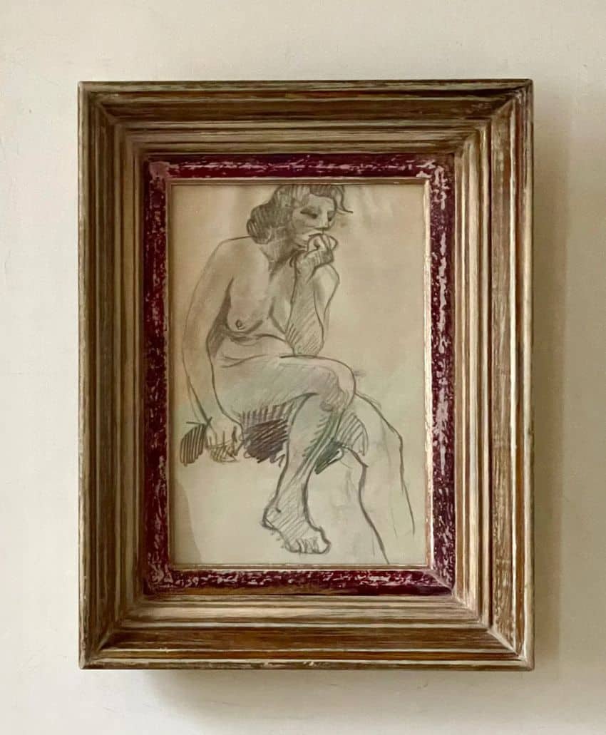 A framed pencil sketch of a seated nude person, posed with one hand resting on their chin and the other holding an object. The frame is wooden with an inner red border, hung on a neutral-colored wall.