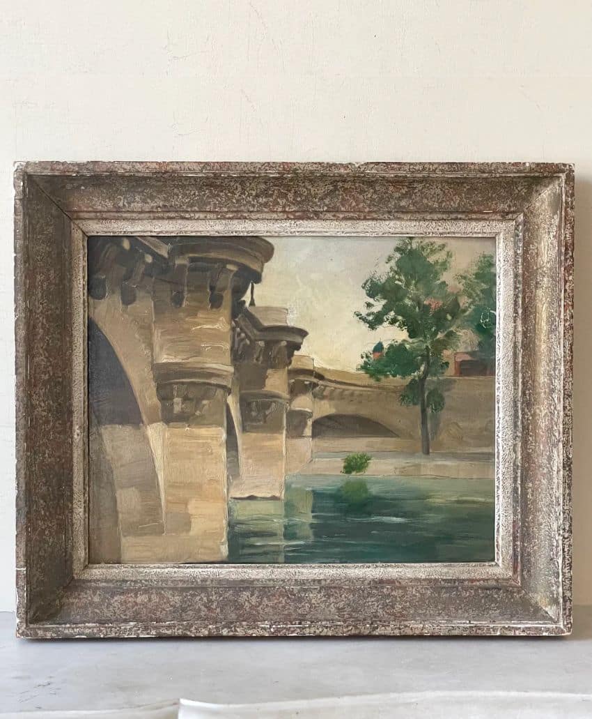Framed painting of a stone bridge over a calm river, with a tree and greenery on the right side. The colors are soft and muted, giving a serene and classical feel to the artwork.