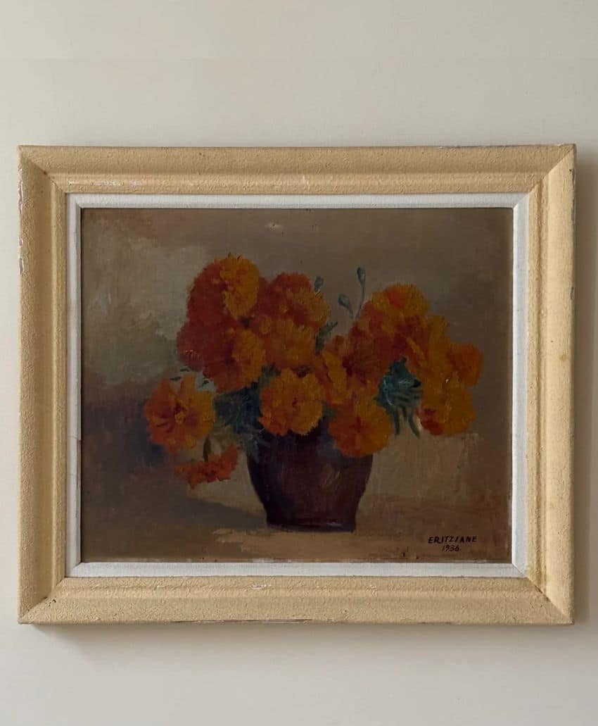 A framed painting of a brown vase filled with vibrant orange and yellow flowers, set against a muted brown background, hanging on a beige wall.