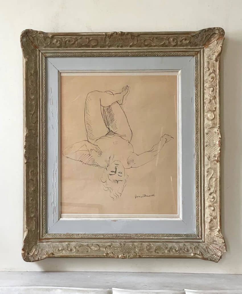 Framed sketch of a nude figure lying on their back with legs raised and arms bent. The drawing is minimalist with simple lines and mounted in an ornate, weathered, gold and gray frame against a plain wall.