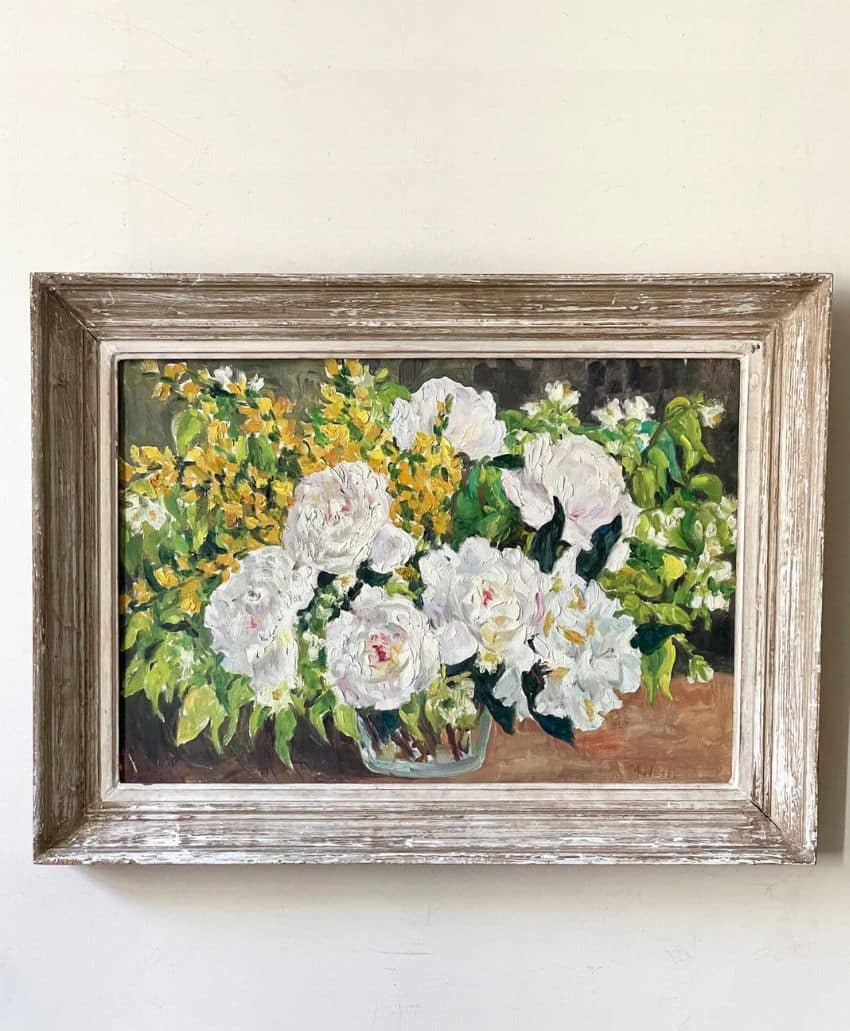 A framed painting of white and yellow flowers with green leaves set against a neutral background, hanging on a plain white wall.