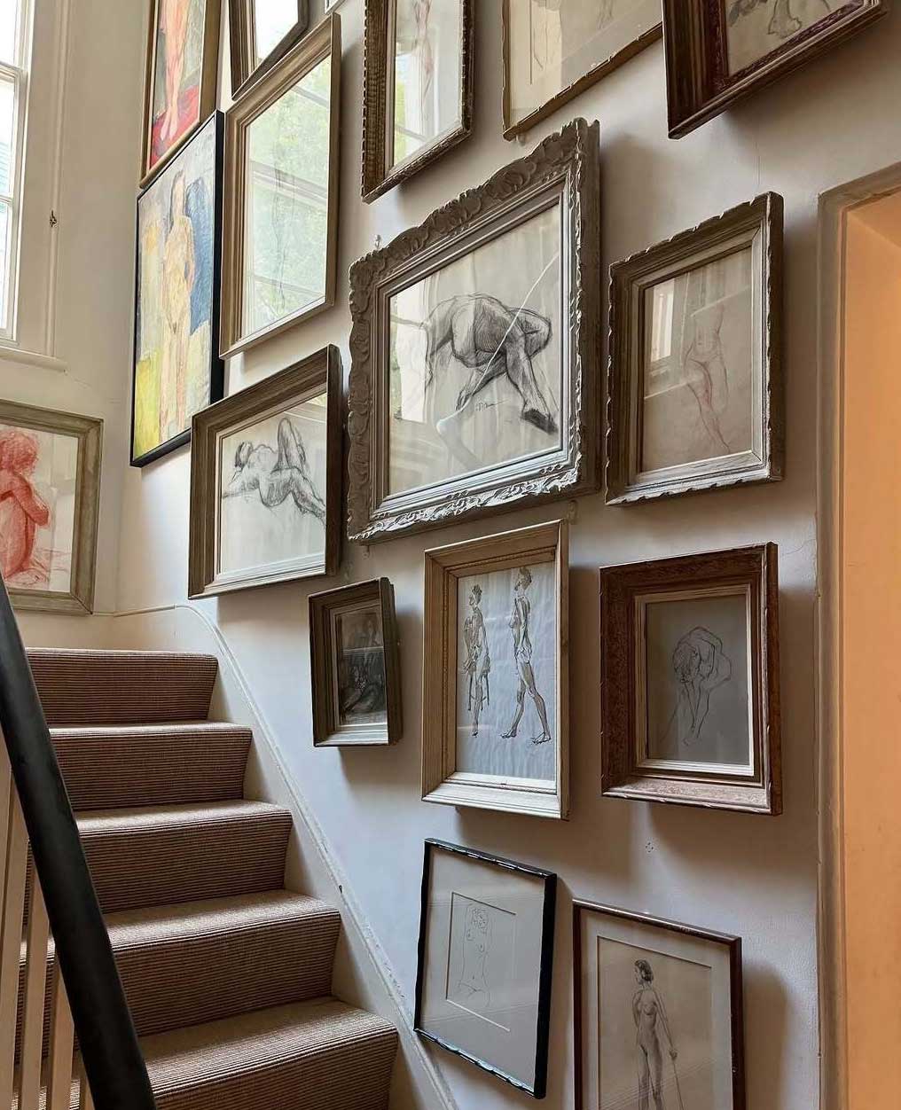 Art-filled staircase with framed sketches and drawings covering the wall above carpeted steps, creating a gallery-like display. Natural light from a nearby window brightens the space.