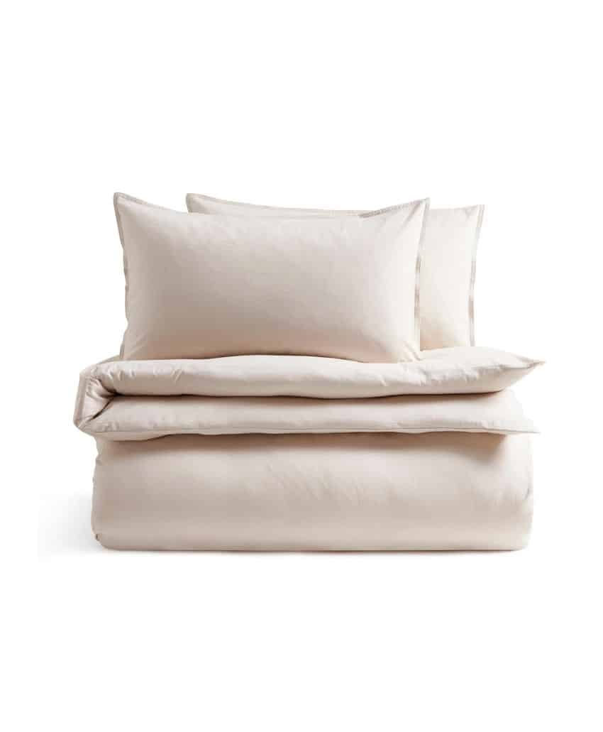 A neatly folded, light beige bedding set with two pillows and a comforter stacked on top of each other, displayed against a white background.