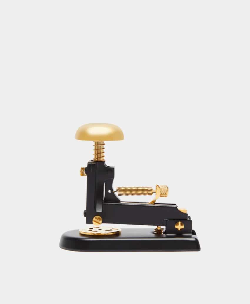A black and gold mechanical device with a round golden button, mounted on a rectangular base, photographed against a plain white background.