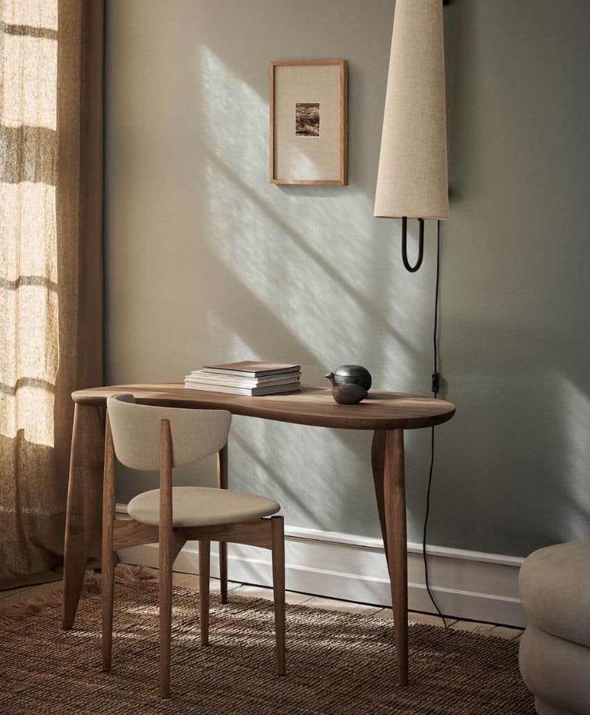 A minimalist wooden desk with a matching chair sits by a window with sheer curtains. On the desk are stacked books and a small decorative object. Soft light casts shadows on the textured wall, where artwork and a lamp hang.