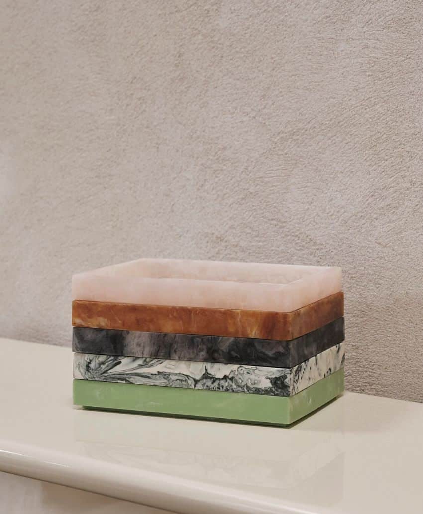 A rectangular box with four stacked layers in different colors: pale pink, brown, marbled black and white, and solid green, set against a textured beige wall on a smooth cream surface.