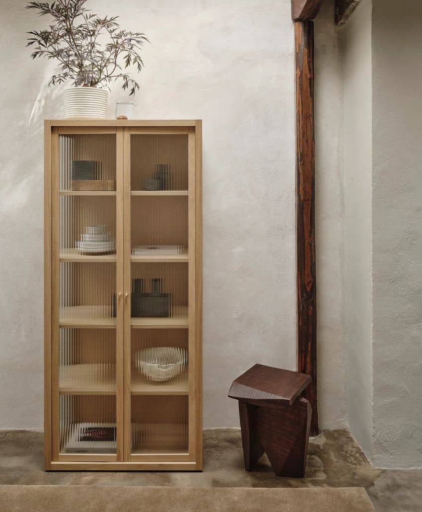 A light wood cabinet with glass doors displaying ceramics stands against a textured beige wall. A potted plant sits on top, and a geometric wooden stool is nearby on a concrete floor.