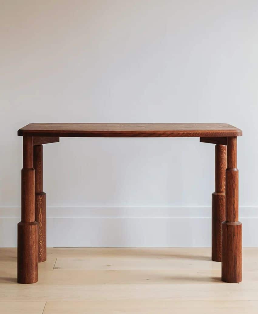 A minimalist wooden table with a rectangular top and four thick, uniquely stacked cylindrical legs, placed against a plain light-colored wall on a light wood floor.