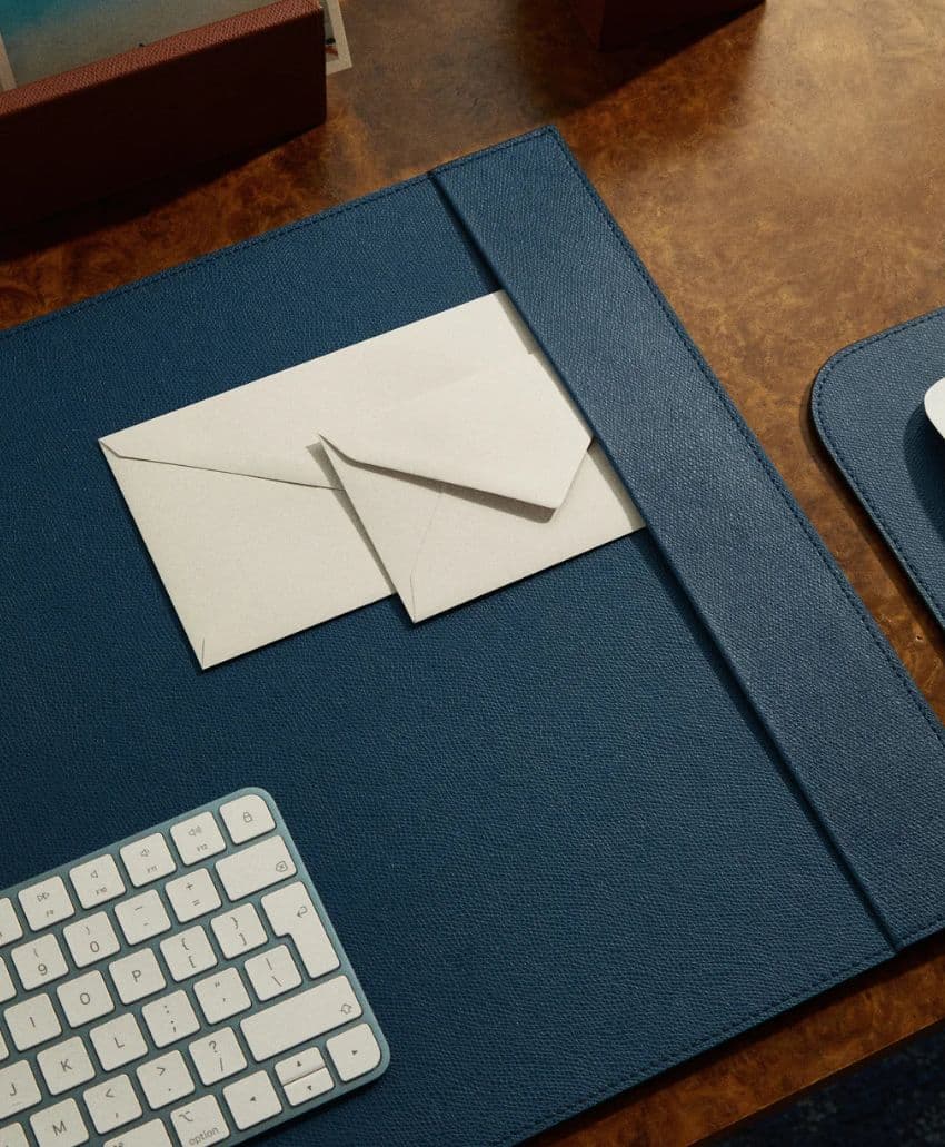 A white keyboard and two white envelopes rest on a blue desk pad atop a brown wooden desk. The scene gives a minimal, organized workspace appearance.