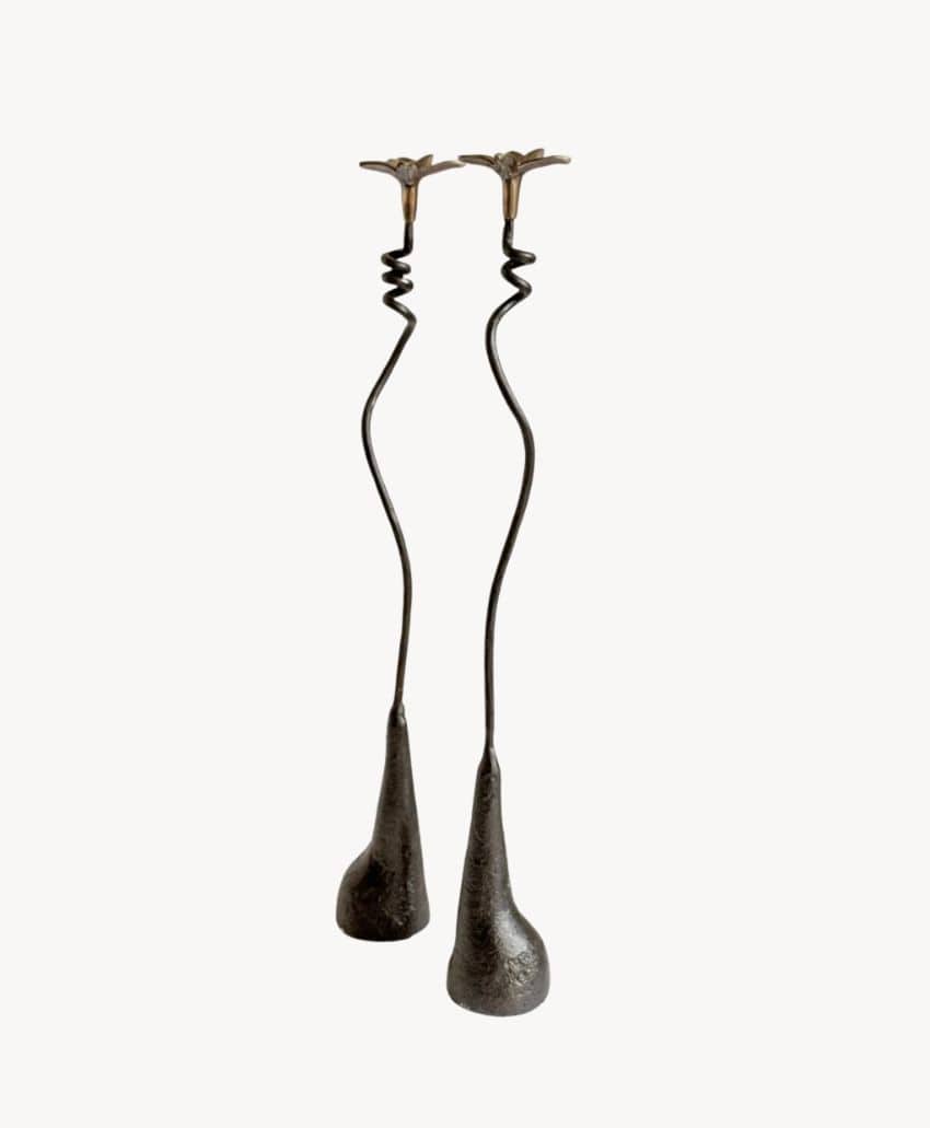 Two tall, black, abstract metal candlestick holders with wavy, slender stems and uneven, textured bases, topped with small flower-like holders, set against a plain white background.