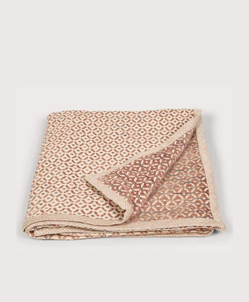 A folded beige and rust-colored blanket with a small geometric pattern, partially flipped at one corner to show both sides, set against a plain light background.