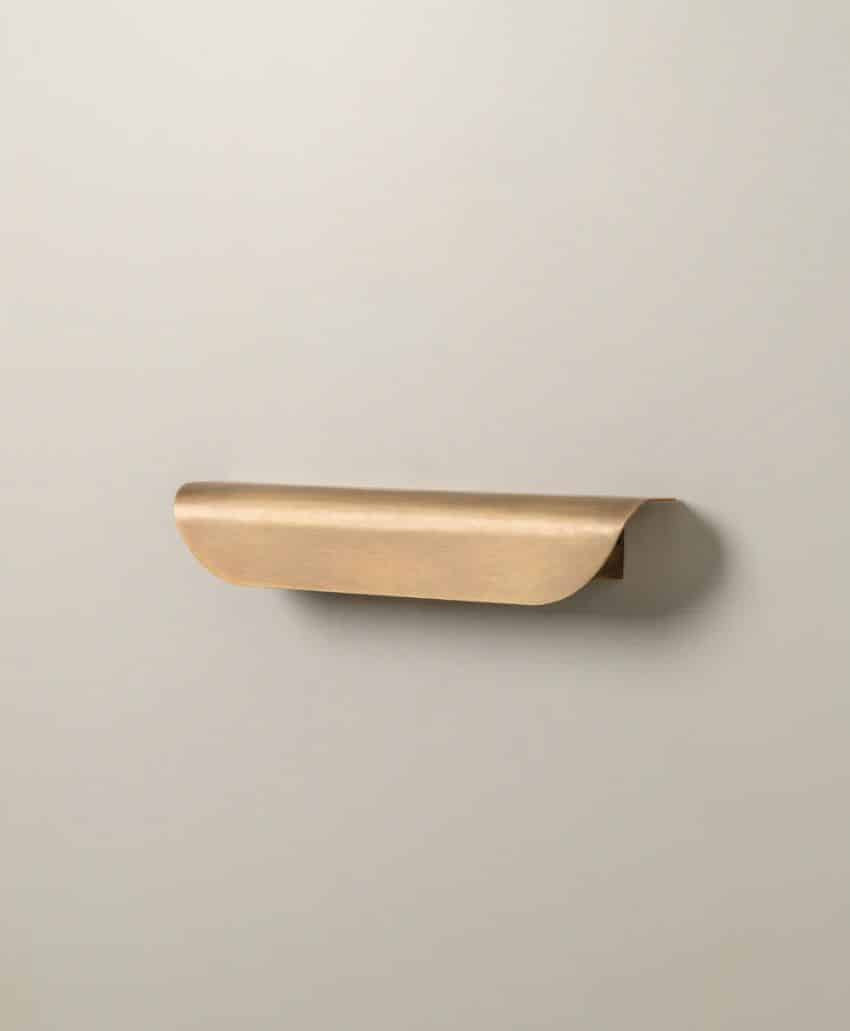 A minimalist brass cabinet handle with a curved design is mounted on a light beige wall.