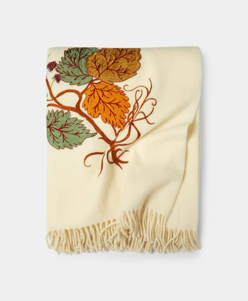 A cream-colored blanket with fringe on the edge, featuring an embroidered design of green, orange, and brown autumn leaves with branches.