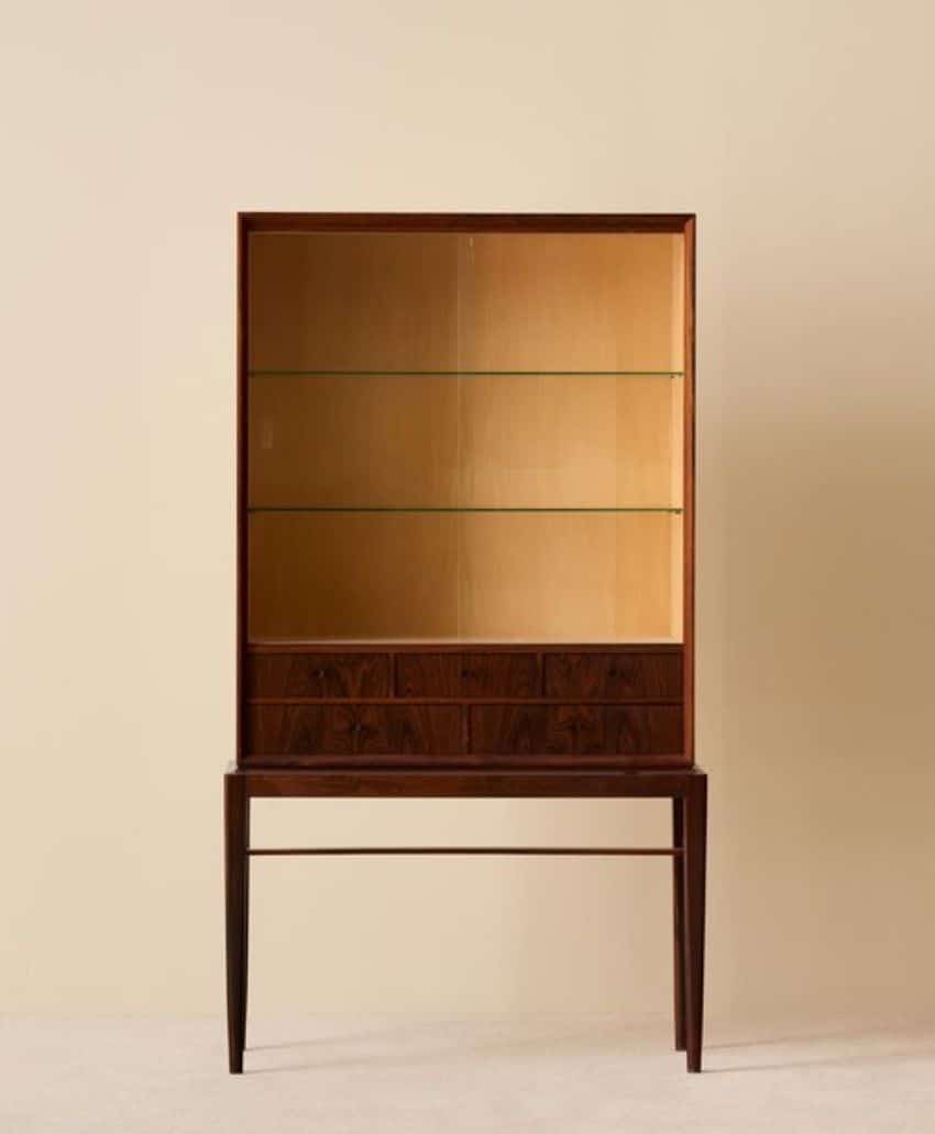 A tall, black, four-door cabinet stands against a light beige wall next to a wooden chair with a black seat and backrest. The floor is light wood.