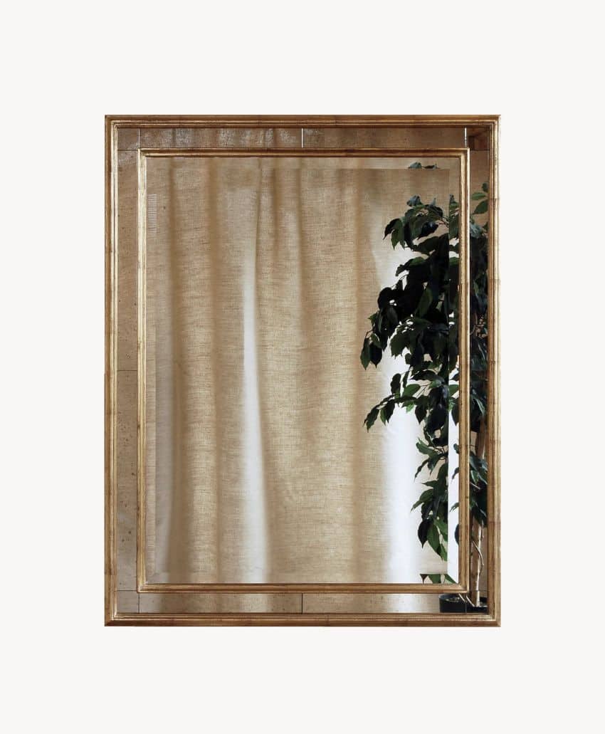 A rectangular mirror with a gold frame reflects beige curtains and part of a leafy green plant on the right side.